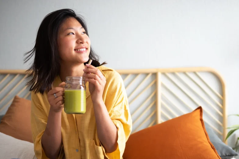 Woman sitting cross-legged on a bed, drinking a green smoothie and smiling