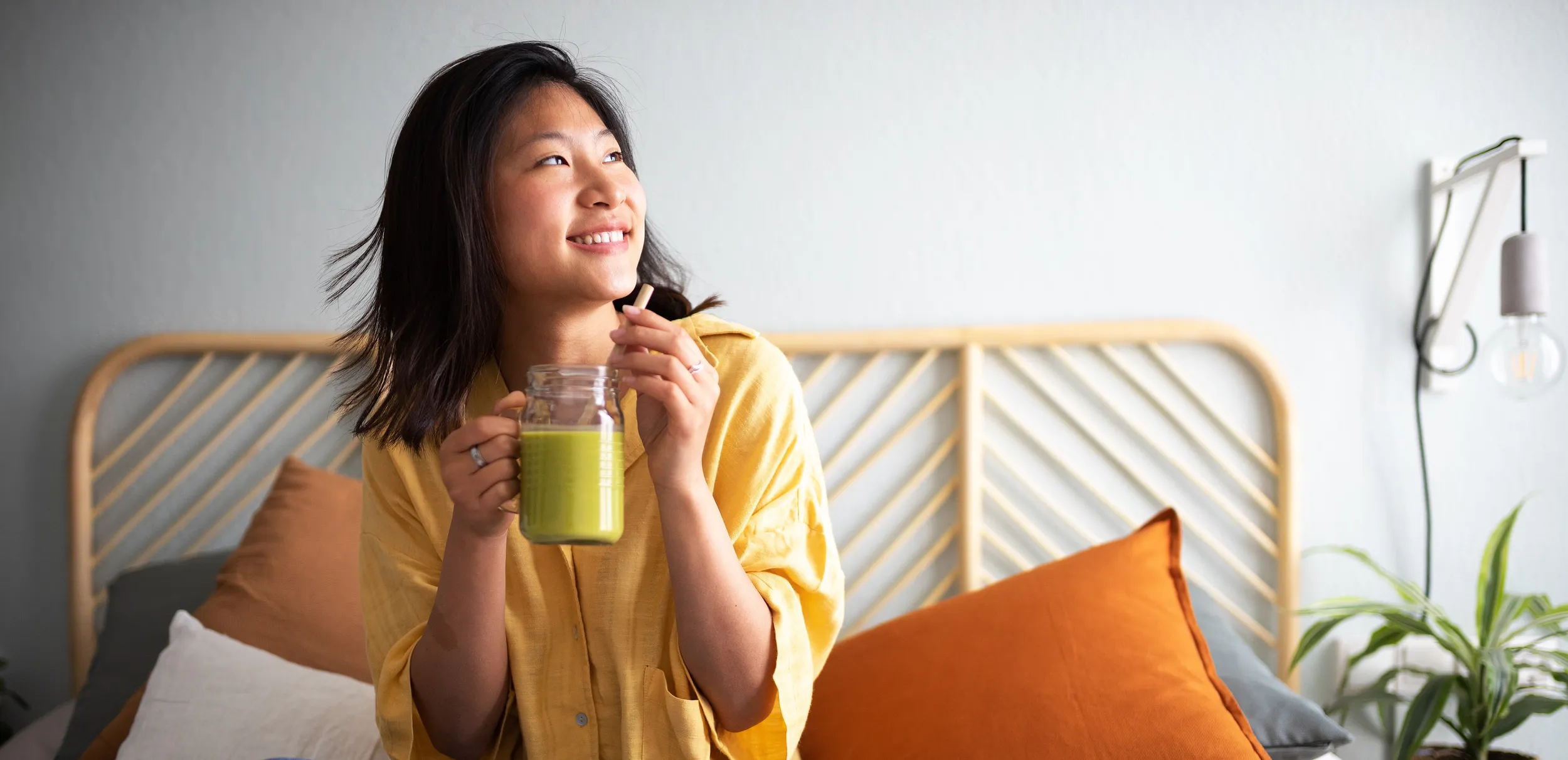 Woman sitting cross-legged on a bed, drinking a green smoothie and smiling