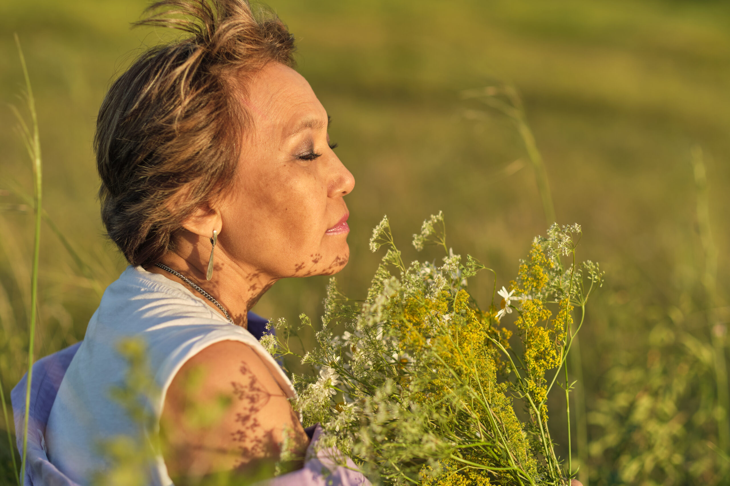 wonderful day: A smiling mature Asian woman enjoys the scent of wildflowers in a sunny park. Her emotions are filled with happiness and joy.