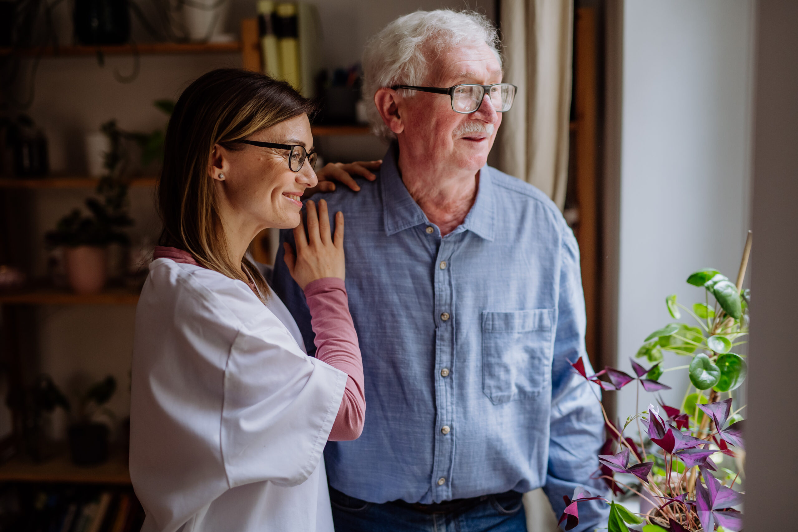 Female caregiver standing beside an elderly man, smiling and offering support as they look out a window together