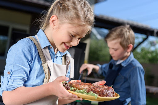 Default ALT text for A young girl thoughtfully looking at a plate of grilled meat and fresh lettuce beside a barbecue grill, illustrating the importance of tailored ketogenic meals in supporting children with autism.