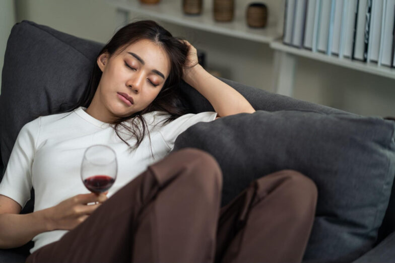 Woman asleep on a couch holding a glass of red wine, suggesting the physical and emotional toll of addiction.