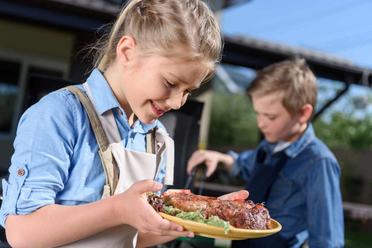 A young girl thoughtfully looking at a plate of grilled meat and fresh lettuce beside a barbecue grill, illustrating the importance of tailored ketogenic meals in supporting children with autism.