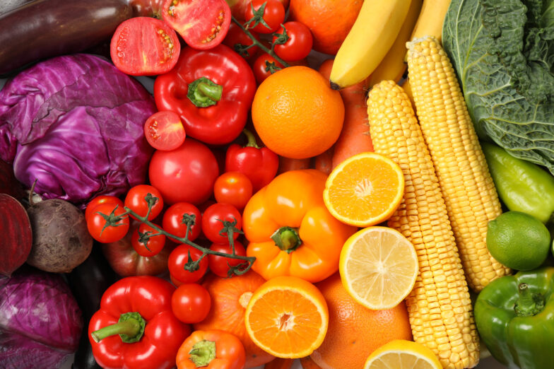 Colorful assortment of fresh fruits and vegetables, including tomatoes, oranges, lemons, bell peppers, corn, cabbage, and bananas, arranged in a vibrant display.