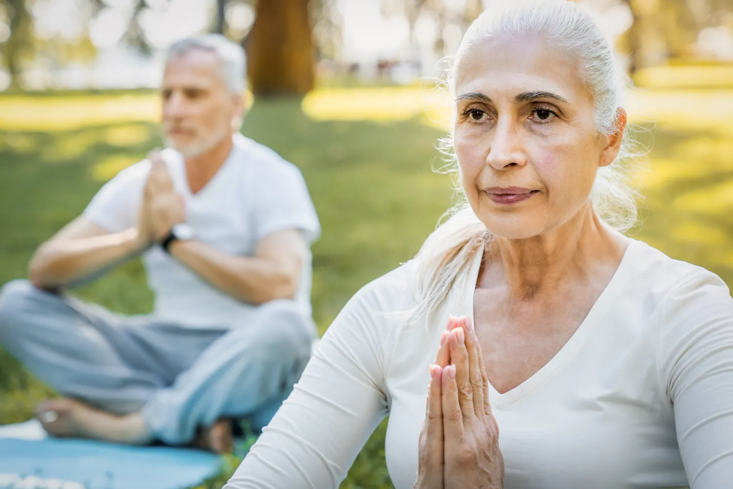 Older woman sitting outdoors in meditation, hands pressed together in prayer position; in the background, an older man appears slightly blurred, also meditating.