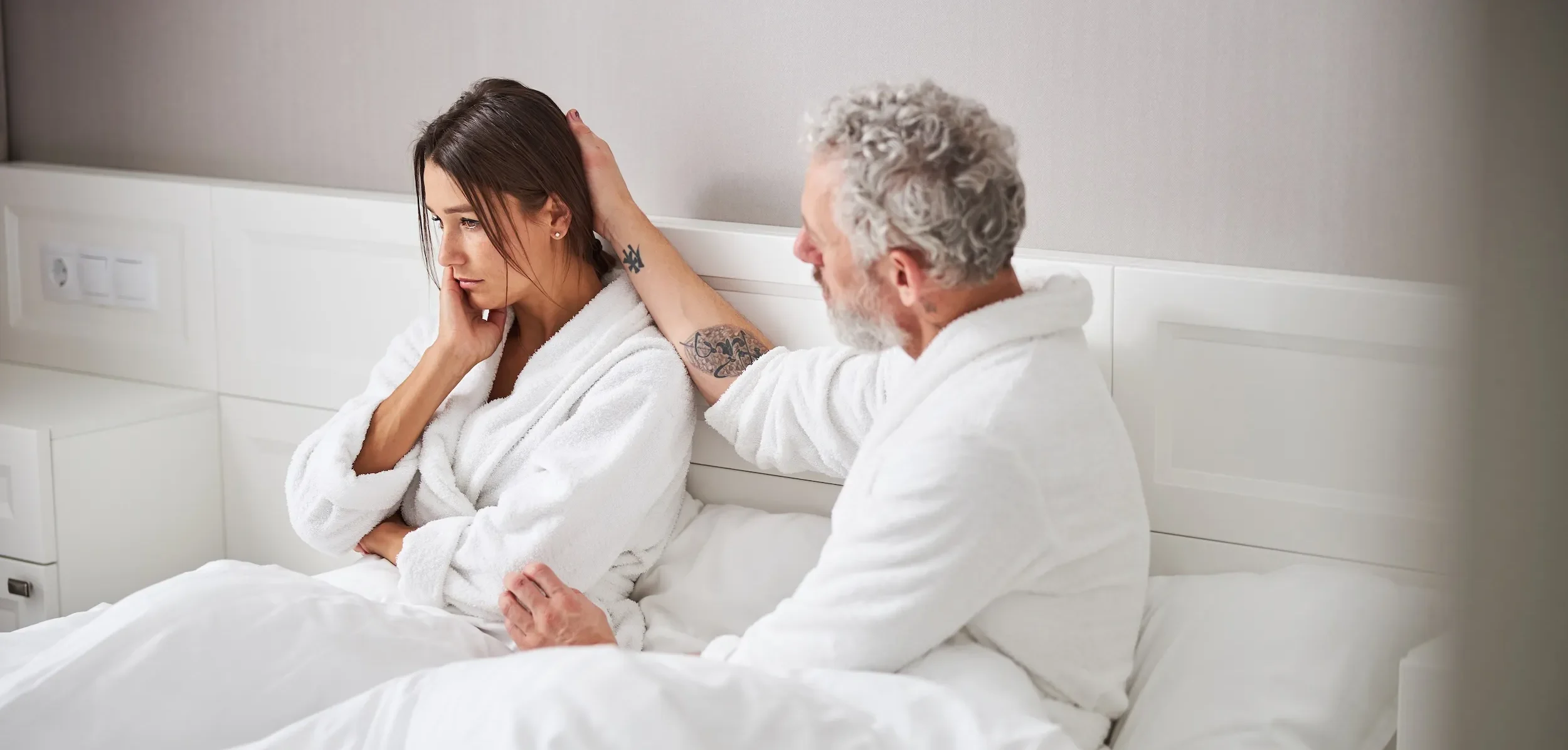 A woman sits on a bed in a white robe, looking away, while a man beside her gently touches her hair.