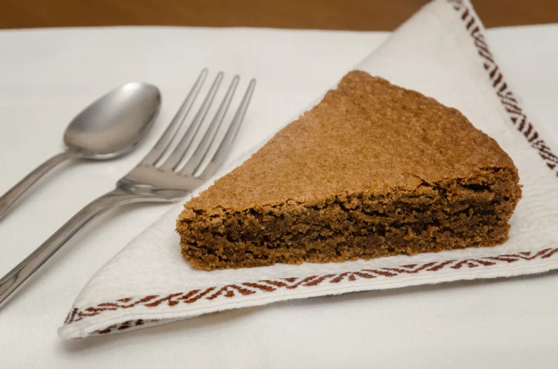 Slice of hazelnut cake on a white napkin with a fork and spoon beside it.
