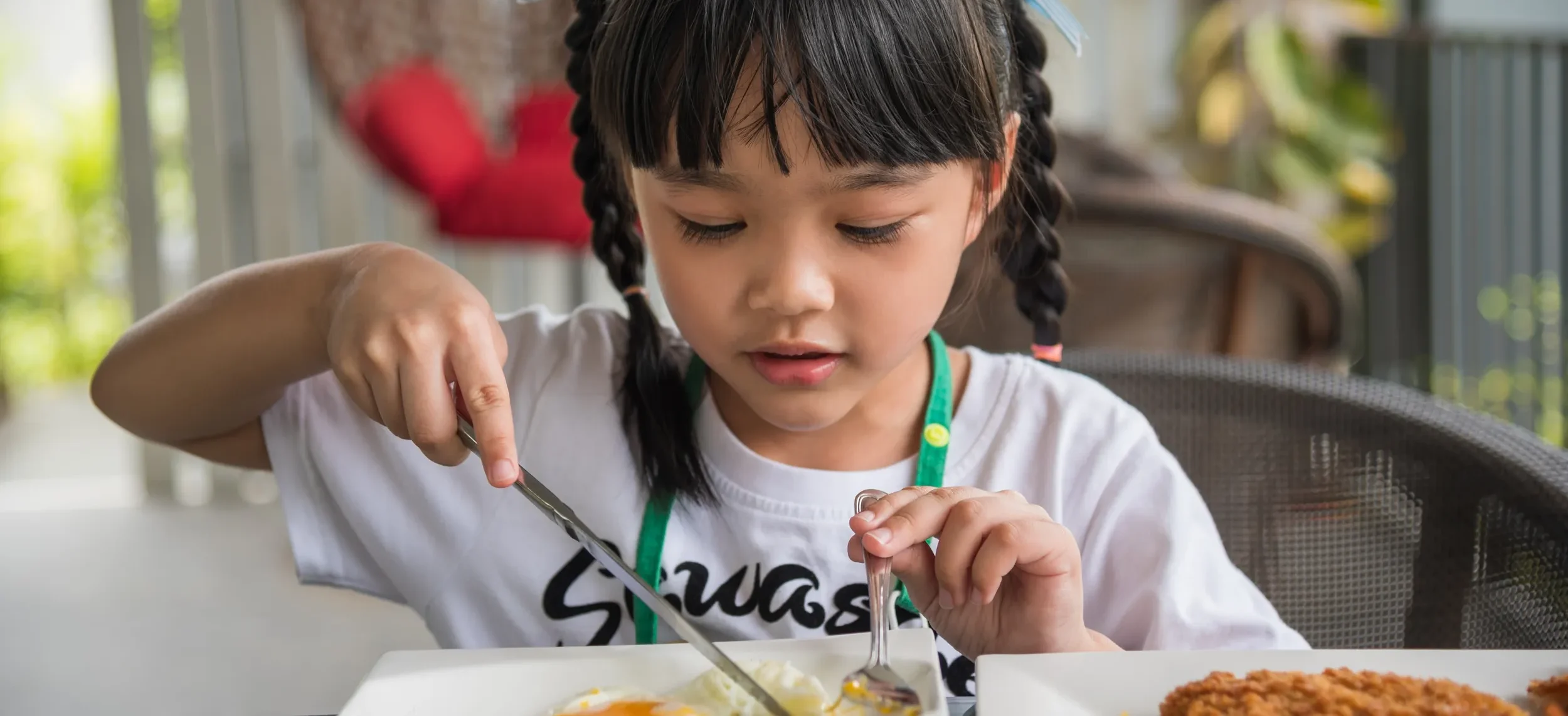 Young girl with braided hair eating a fried egg at a table, focusing carefully on her plate.