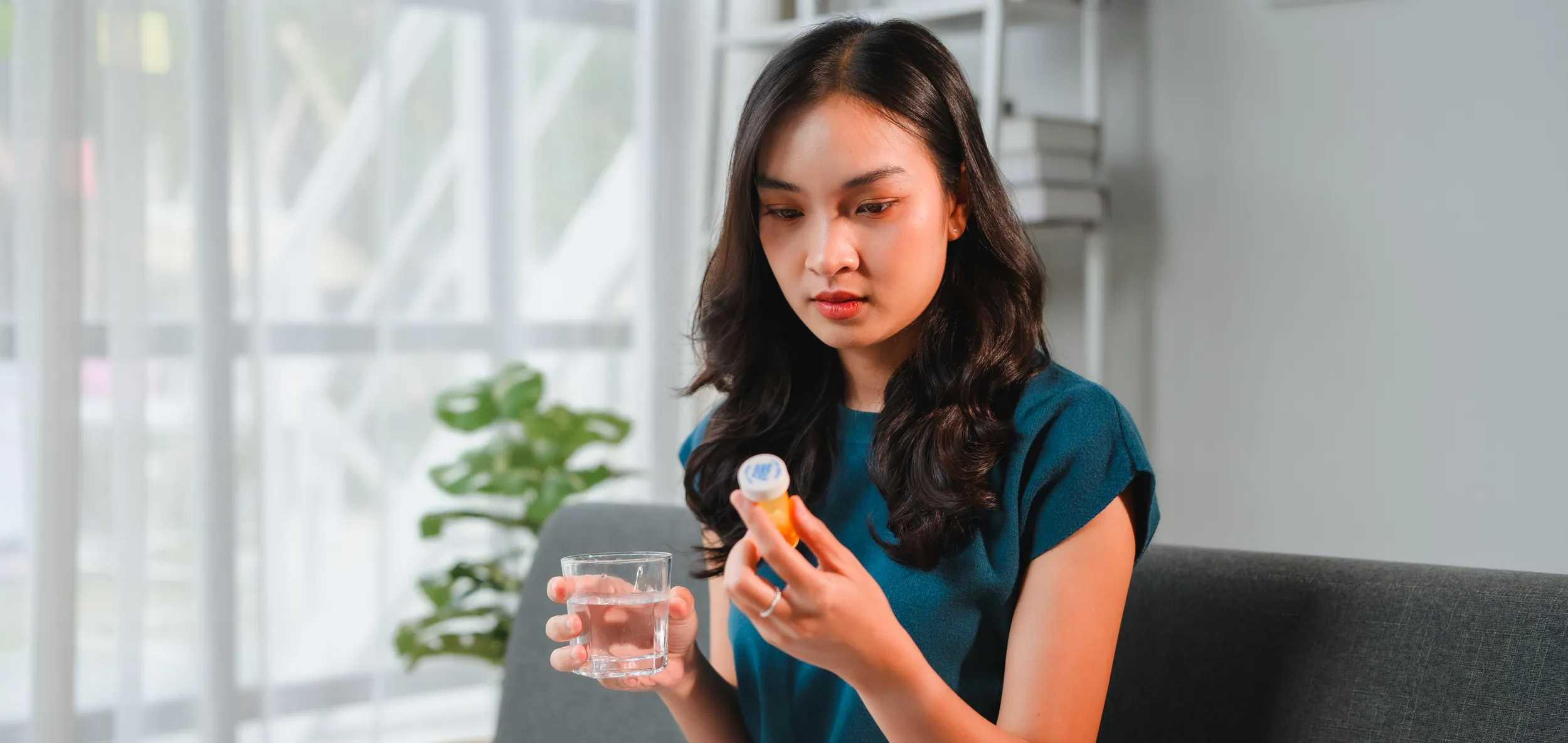 Woman sitting on a couch holding a prescription medication bottle and a glass of water.