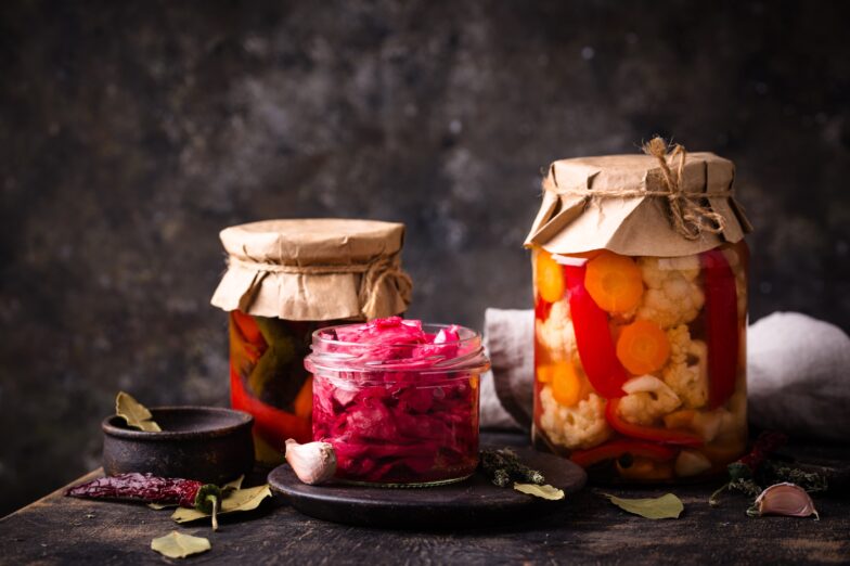 Glass jars of colorful fermented vegetables, including cabbage, carrots, cauliflower, and peppers, arranged on a dark surface with garlic and herbs.
